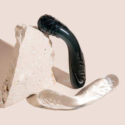 Bent thick glass pleasure wand pictured in two colour ways - midnight black and clear. Thicker ends with ridges and bent in the middle. Pictured on a beige background with stone prop.