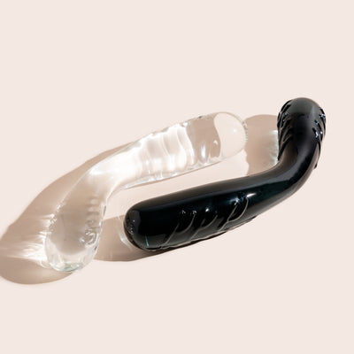 Bent thick glass pleasure wand pictured in two colour ways - midnight black and clear. Thicker ends with ridges and bent in the middle. Pictured on a beige background.