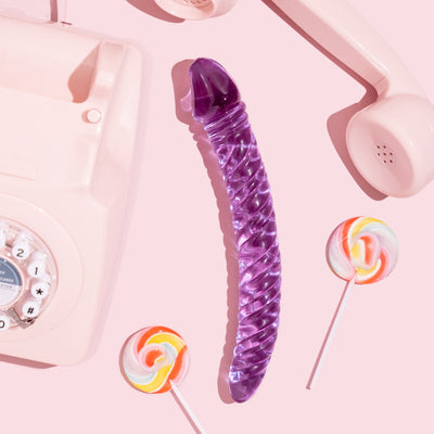 Curved purple glass ridged pleasure wand with thicker Fallic head and tapered end.  Pictured on pink background with lollipops and a pink retro dial telephone.